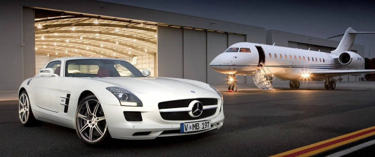 A sleek white Mercedes-Benz sports car parked on an airport tarmac in front of a private jet with its door open and stairs extended. The backdrop includes a large, illuminated hangar with an open door, showcasing the luxurious ambiance of the scene. The photo is taken at dusk or nighttime, adding a dramatic effect with the jet and car both highlighted by soft lighting.